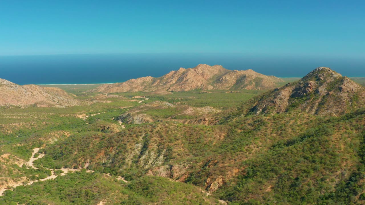 El Salado Viejo Los Cabos aerial frame showing a darker foreground ridge, lighter tan mountains beyond it, green terrain below, blue ocean in back, and no visible people.