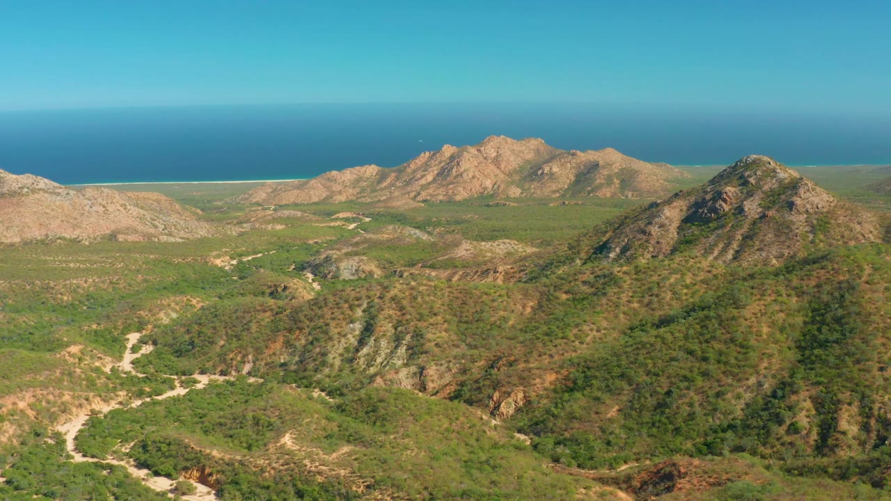 Aerial frame from El Salado Viejo Los Cabos showing green foreground hills, pale dirt tracks through the valley, rocky mountains, a blue ocean horizon, and no visible people.