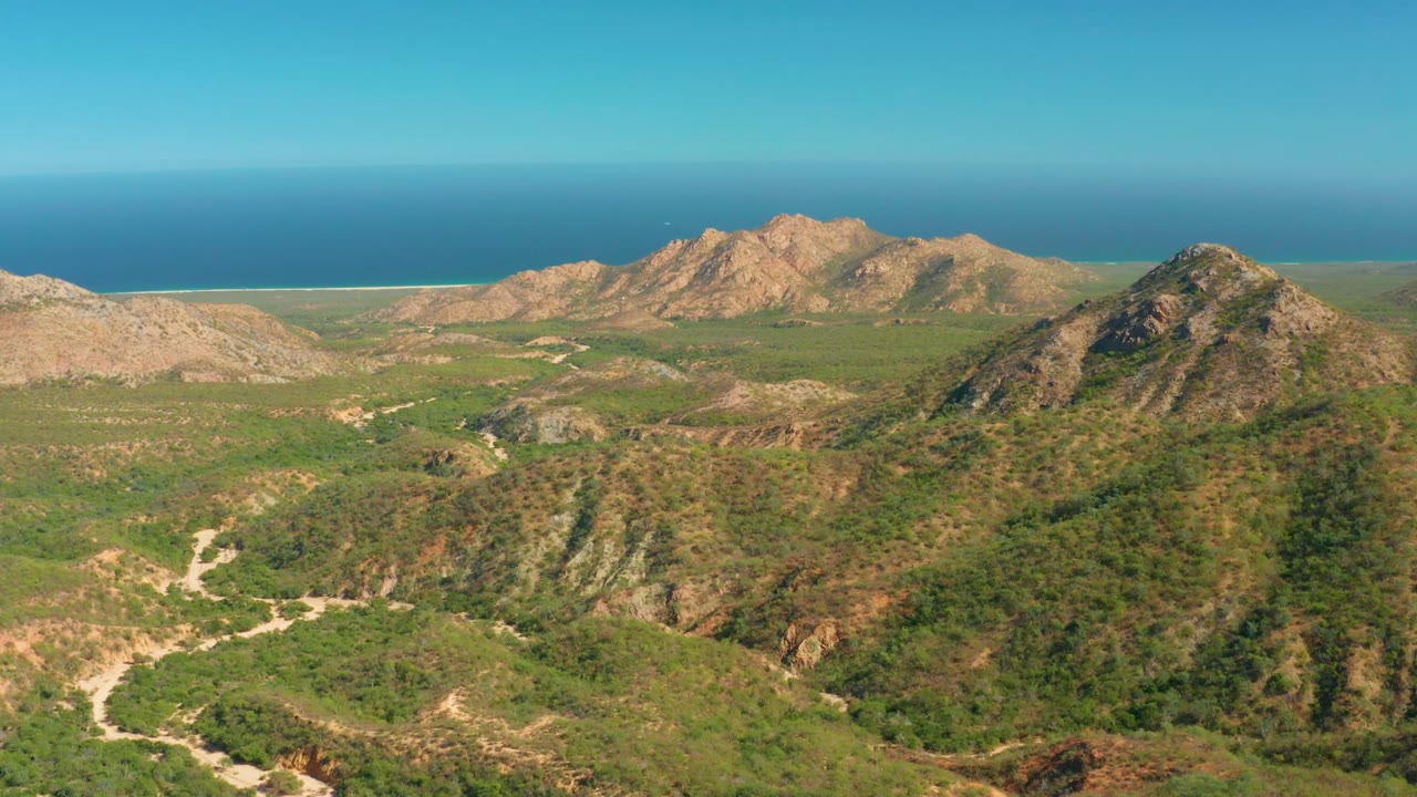 Wide aerial landscape with scrubby hills, winding dirt road, tan mountains, ocean, and clear sky.