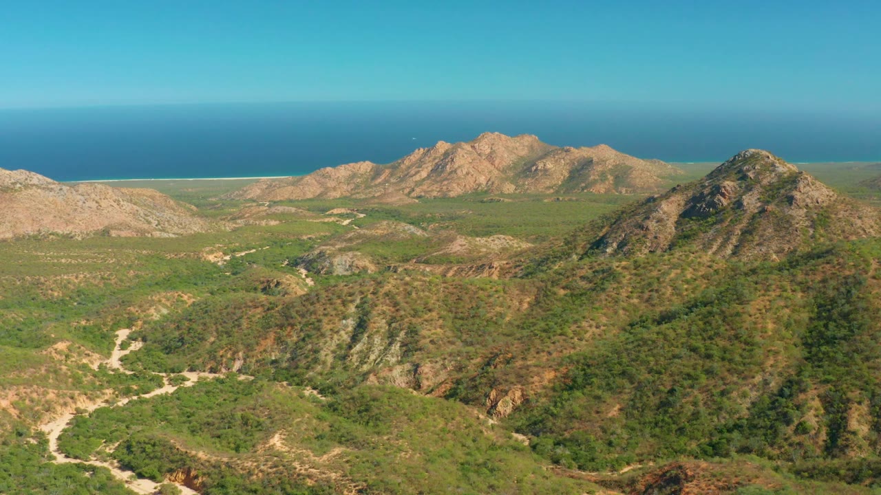 Aerial frame from El Salado Viejo Los Cabos showing a broad green valley, pale road lines across the lower slopes, two rocky ridges in the distance, the ocean horizon, and no visible people.