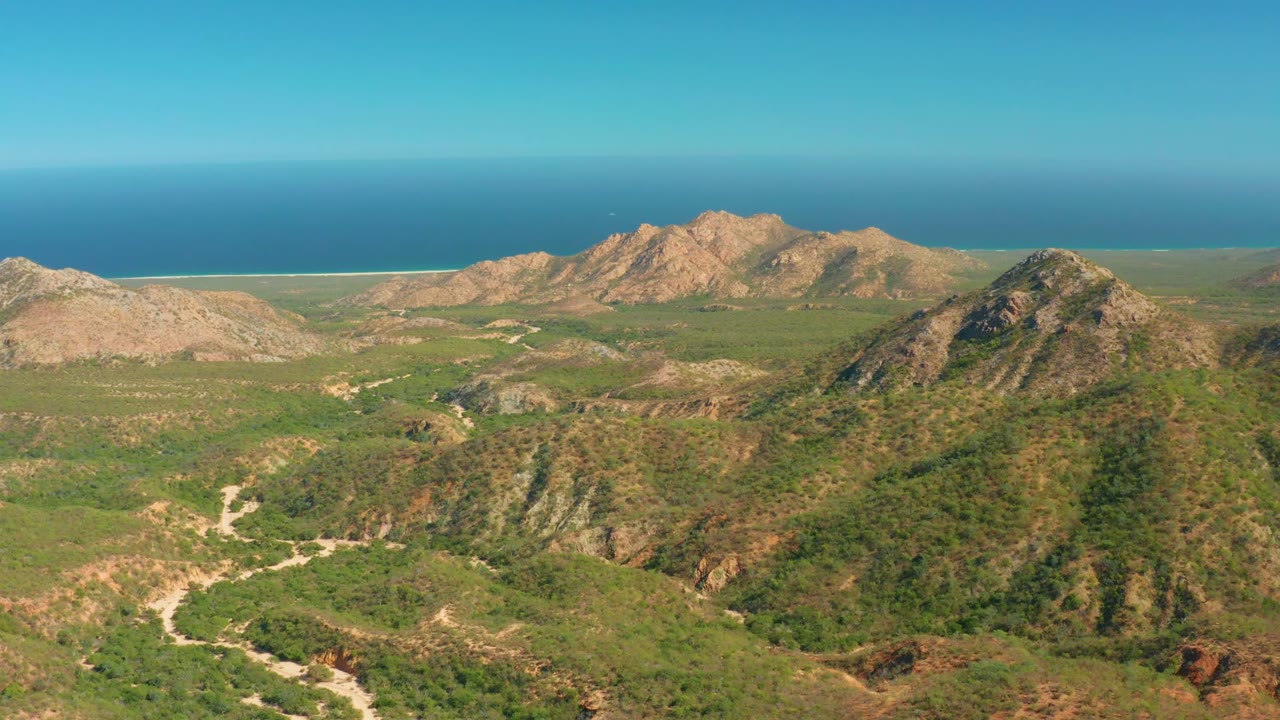 Opening aerial frame over El Salado Viejo Los Cabos, with green hills and pale winding roads below rocky peaks, ocean on the horizon, and no visible people.