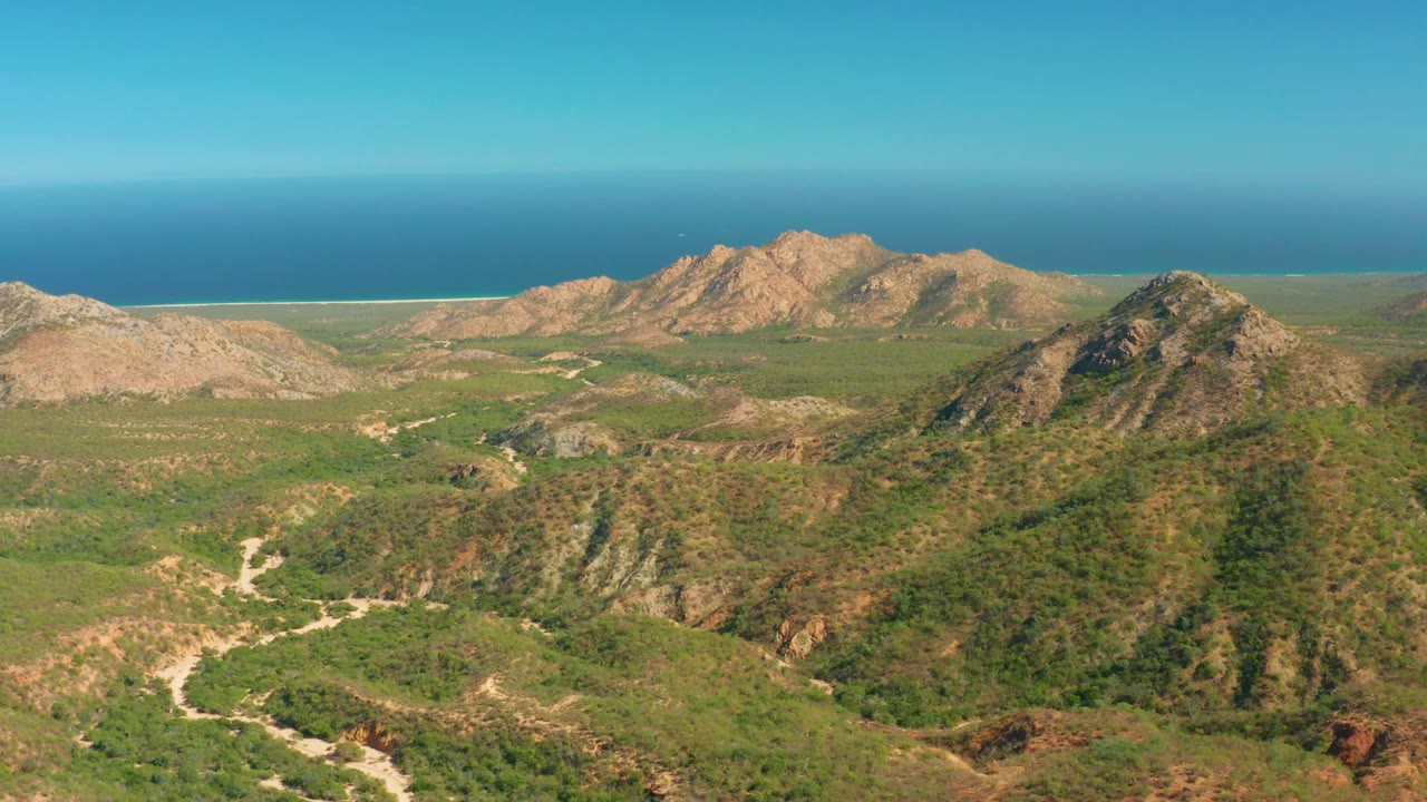 Coastal mountain valley with green slopes, rocky ridges, dirt roads, and blue ocean beyond.