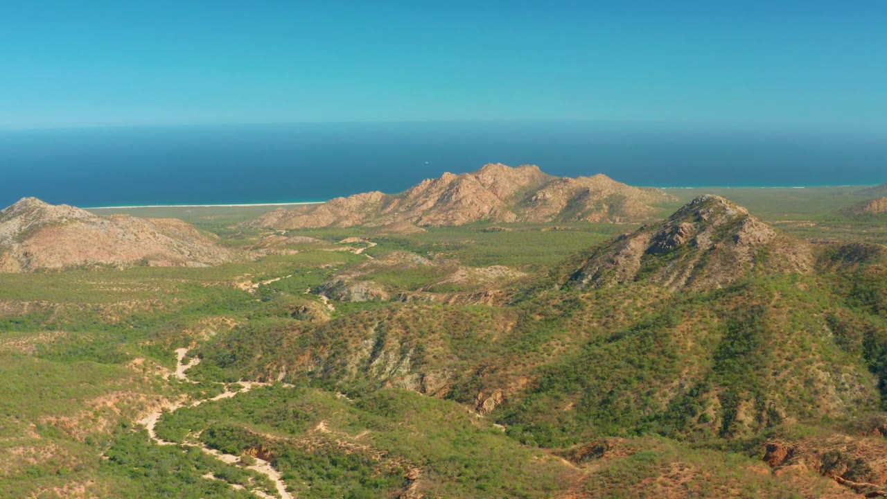 Opening frame over El Salado Viejo Los Cabos, with scrub-covered hills in front, tan rocky ridges behind, a blue ocean horizon, clear sky, and no visible people.