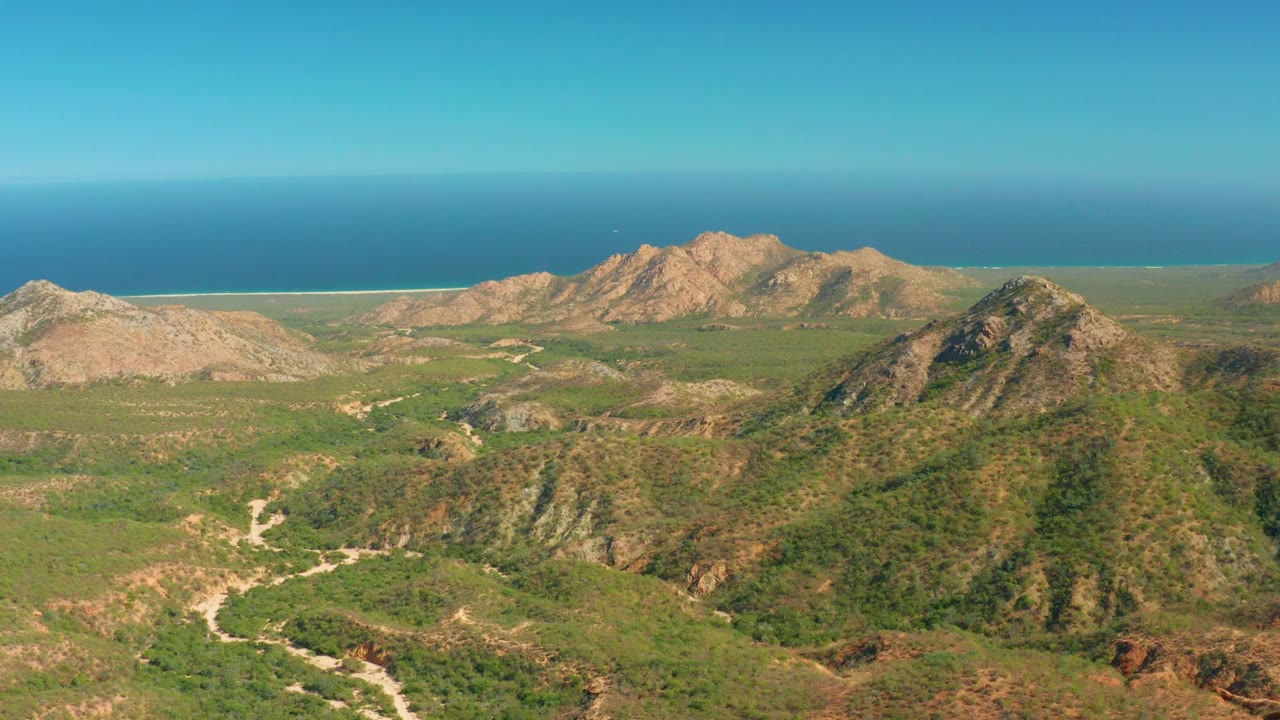 Aerial coastal terrain with green slopes, tan mountains, pale tracks, ocean horizon, and clear sky.