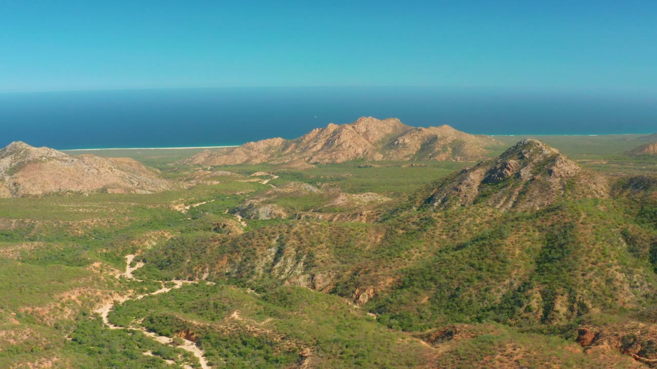 At 0:02, the aerial composition shows El Salado Viejo Los Cabos hills filling the lower frame, layered rocky ridges across the middle, ocean at the horizon, and no visible people.