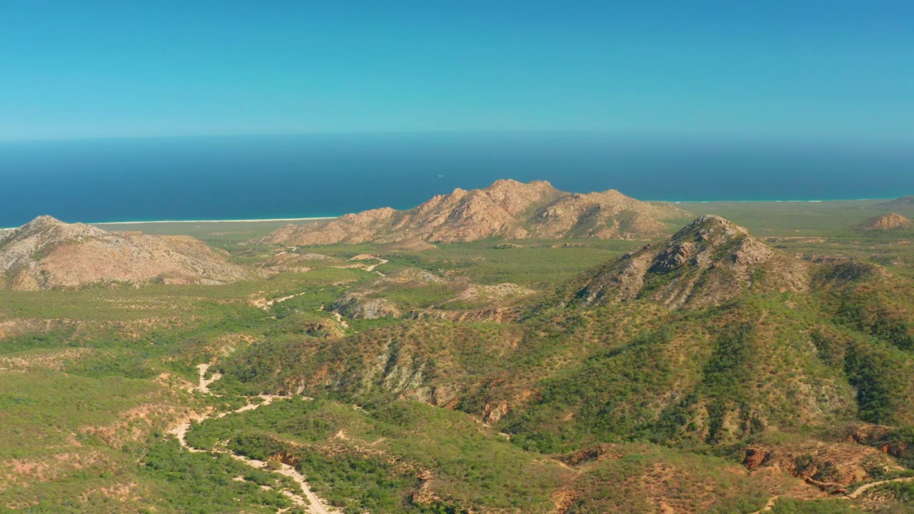 At 0:00, an aerial view over El Salado Viejo Los Cabos shows green hills, rocky peaks, pale winding dirt roads, a distant blue ocean horizon, and no visible people.