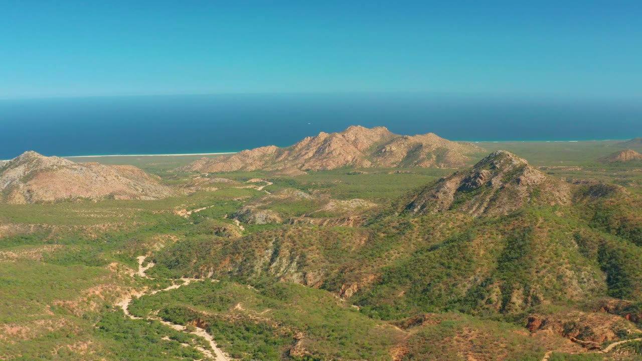Wide aerial landscape with rocky ridges, green valleys, pale road tracks, and ocean horizon.