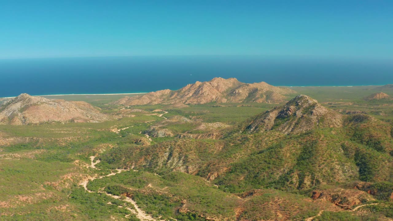 Opening frame of El Salado Viejo Los Cabos showing green rocky hills, a pale winding road through the valley, tan ridges, blue ocean, clear sky, and no visible people.