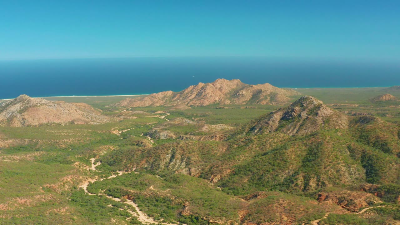 At 0:02, the static aerial view over El Salado Viejo Los Cabos shows the foreground ridge, winding pale road, broad green valley, rocky tan peaks, blue ocean band, and no visible people.