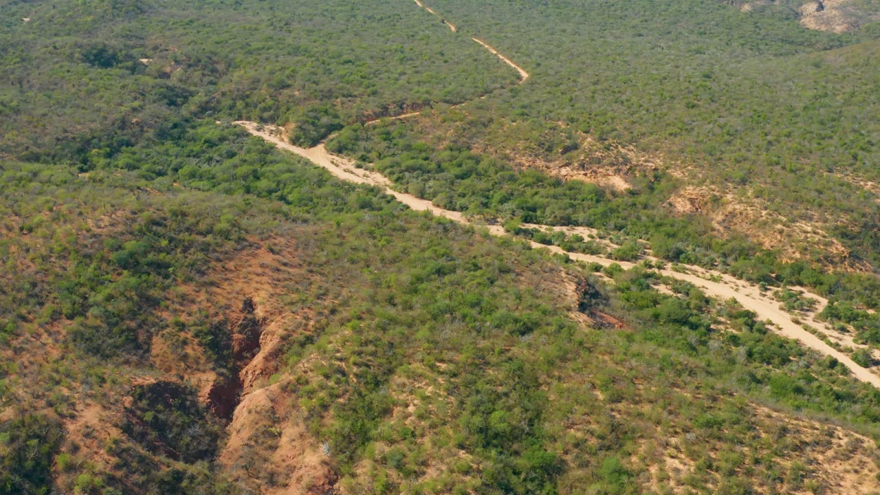 Aerial frame over El Salado Viejo Los Cabos with a pale dirt road bending through green scrub, reddish exposed slopes, and no visible people.