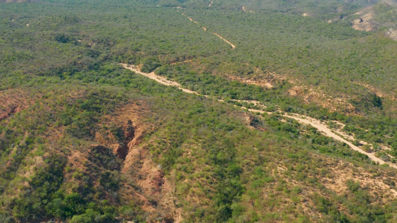 Aerial frame over El Salado Viejo Los Cabos showing a pale dirt road curving through green hills and exposed reddish slopes, with no visible people.