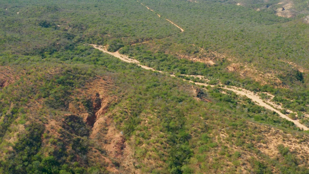 High aerial frame over El Salado Viejo Los Cabos showing the road bending across a valley floor between green hills and reddish exposed ravines, with no visible people.