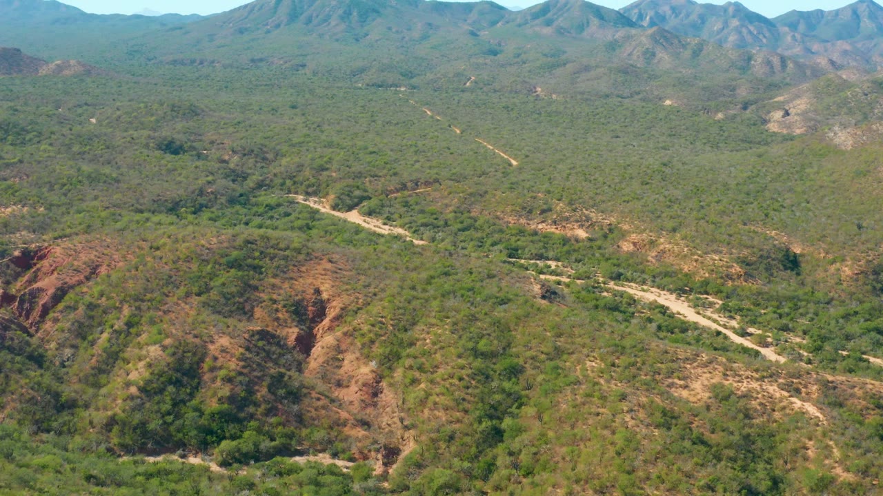 Aerial frame over El Salado Viejo, Los Cabos, showing a pale dirt road crossing green hilly terrain toward distant mountains, with no visible people.