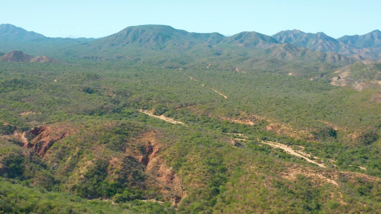 Opening frame of El Salado Viejo Los Cabos with scrub-covered hills, a pale dirt road crossing the terrain, distant mountain ridges, and no visible people.