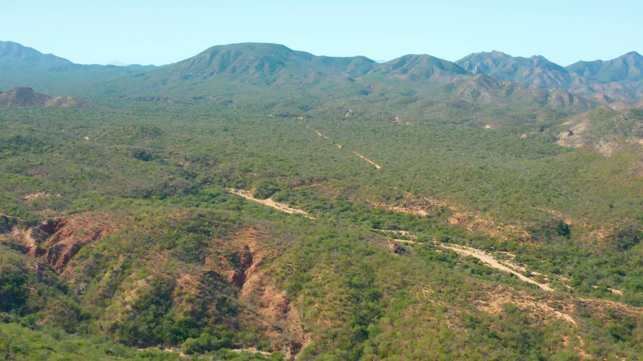 Midwide El Salado Viejo Los Cabos view with reddish eroded slopes in the lower frame, a pale road bending through green vegetation, distant peaks, and no visible people.