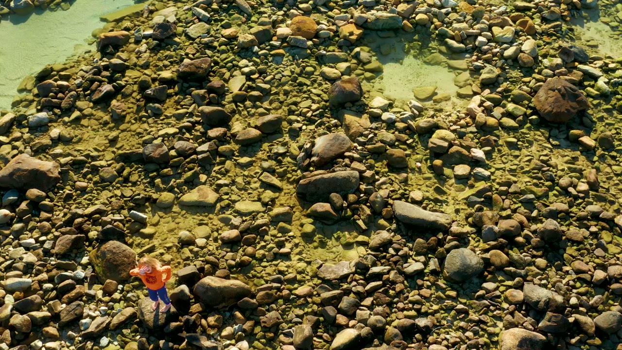 At 0:00, an overhead view in El Tule, Los Cabos shows a person in an orange shirt standing among rounded river rocks beside shallow green water.