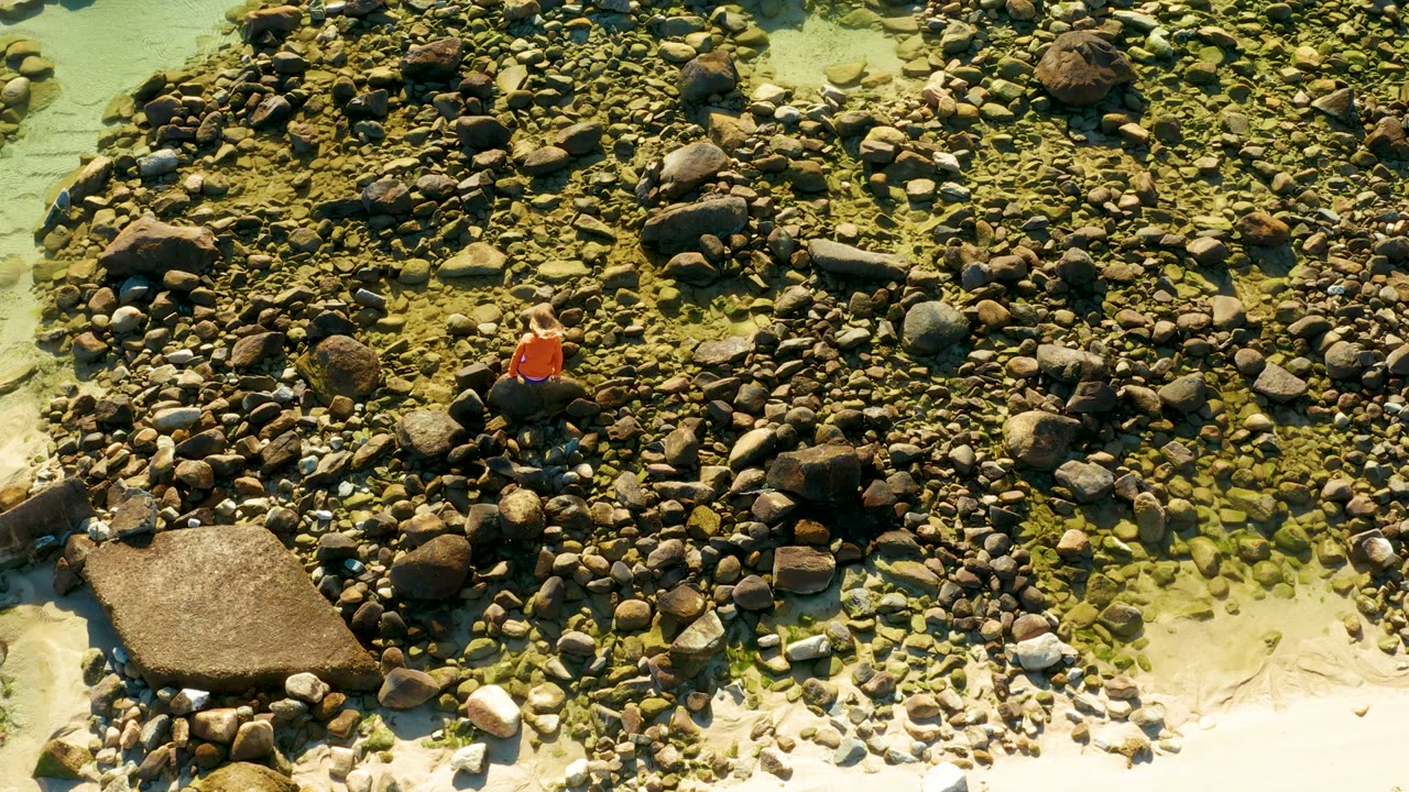 Overhead frame from El Tule, Los Cabos shows a person in an orange shirt near the upper left area of a rocky shore with pale sand and clear shallow water below.