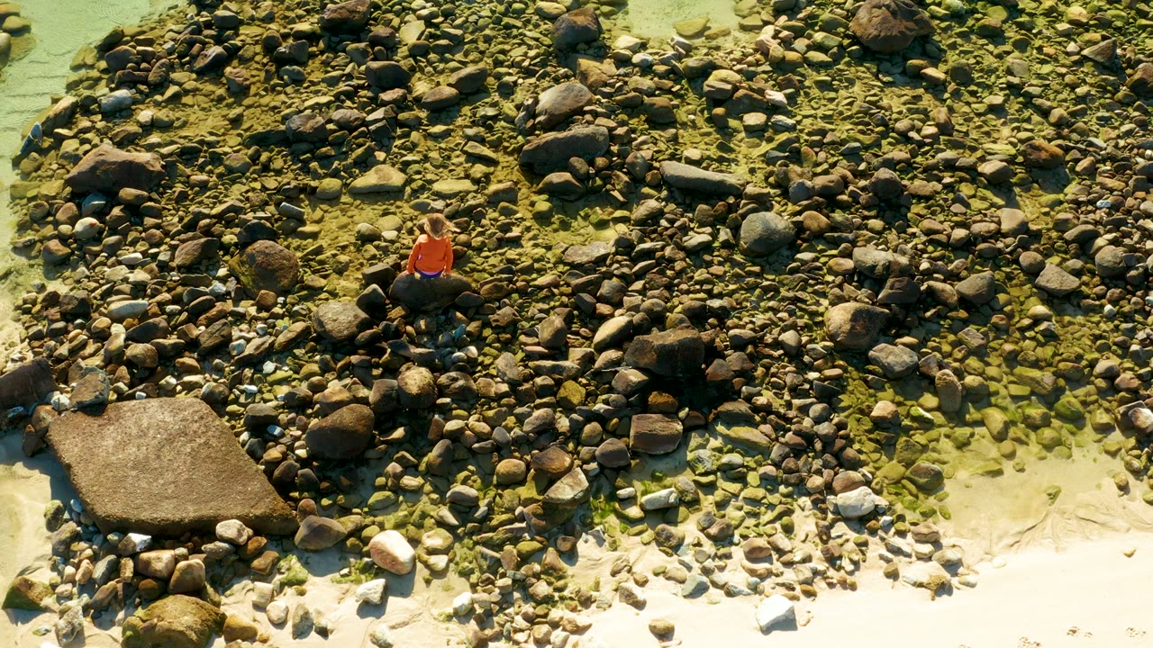 Person in orange shirt on a boulder above sand and shallow water.