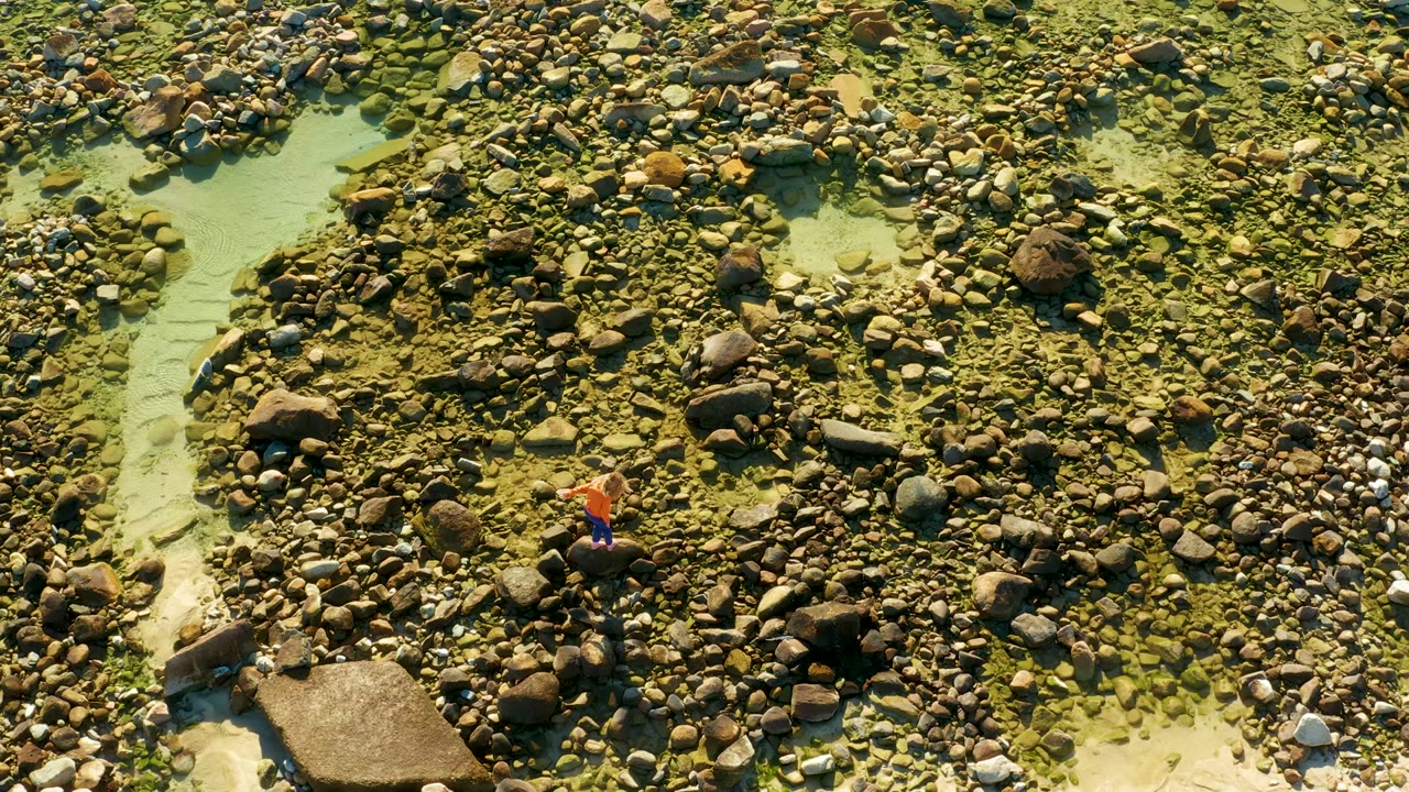 Overhead view in El Tule Los Cabos of a person in an orange shirt stepping among rounded rocks beside a pale channel of clear shallow water.