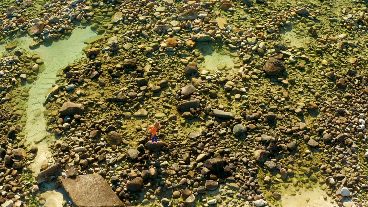 Overhead El Tule Los Cabos frame showing the person slightly farther across the stones, with clear water cutting through the rocks on the left.