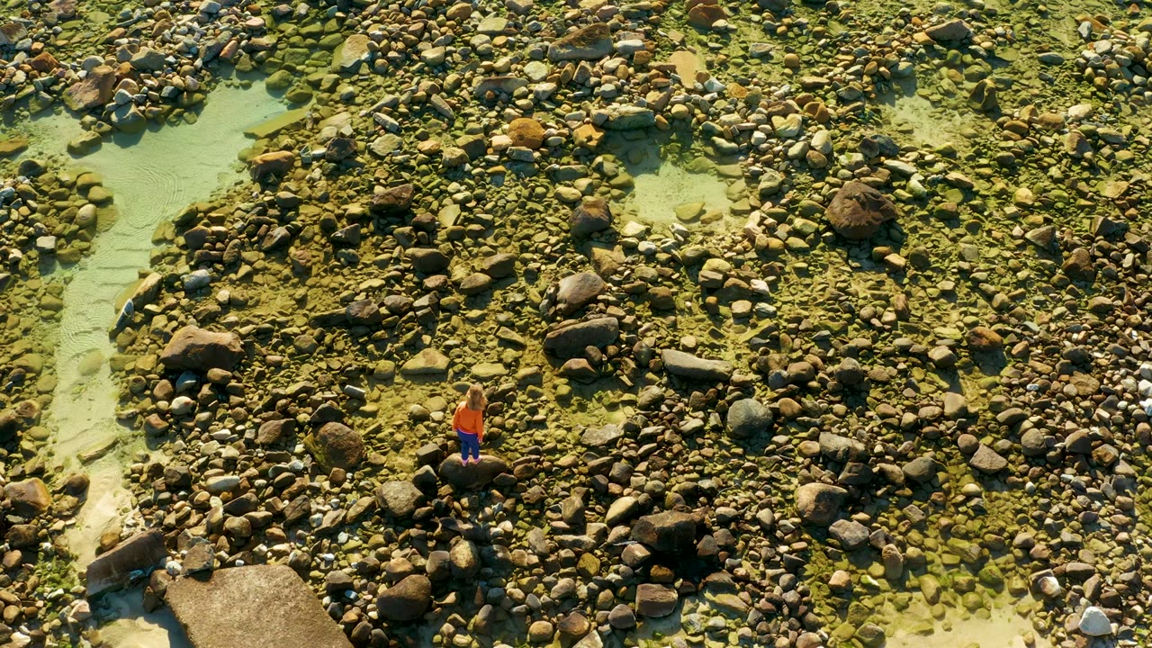 Overhead view in El Tule Los Cabos with the person centered among dense rounded stones, pale green water, and a broad flat rock near the lower left.