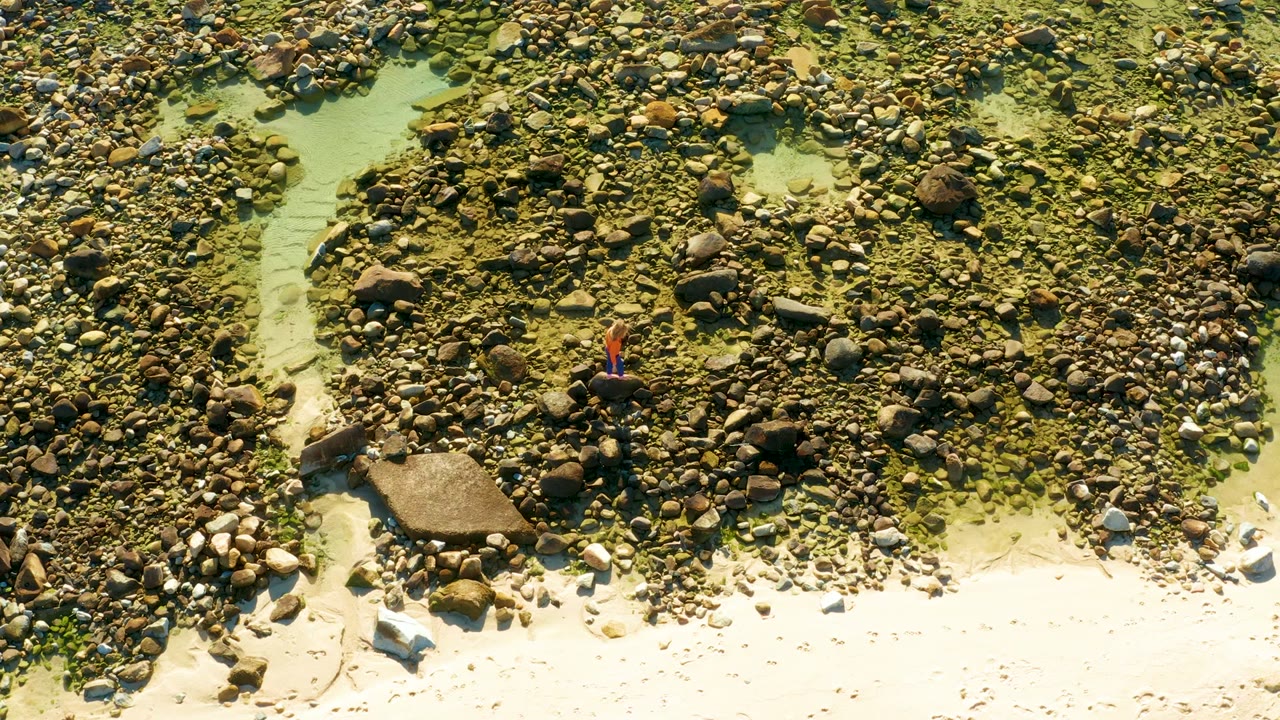 Overhead frame from El Tule Los Cabos showing a person in a red shirt near the left side of a rocky shallow stream with pale sand along the bottom edge.