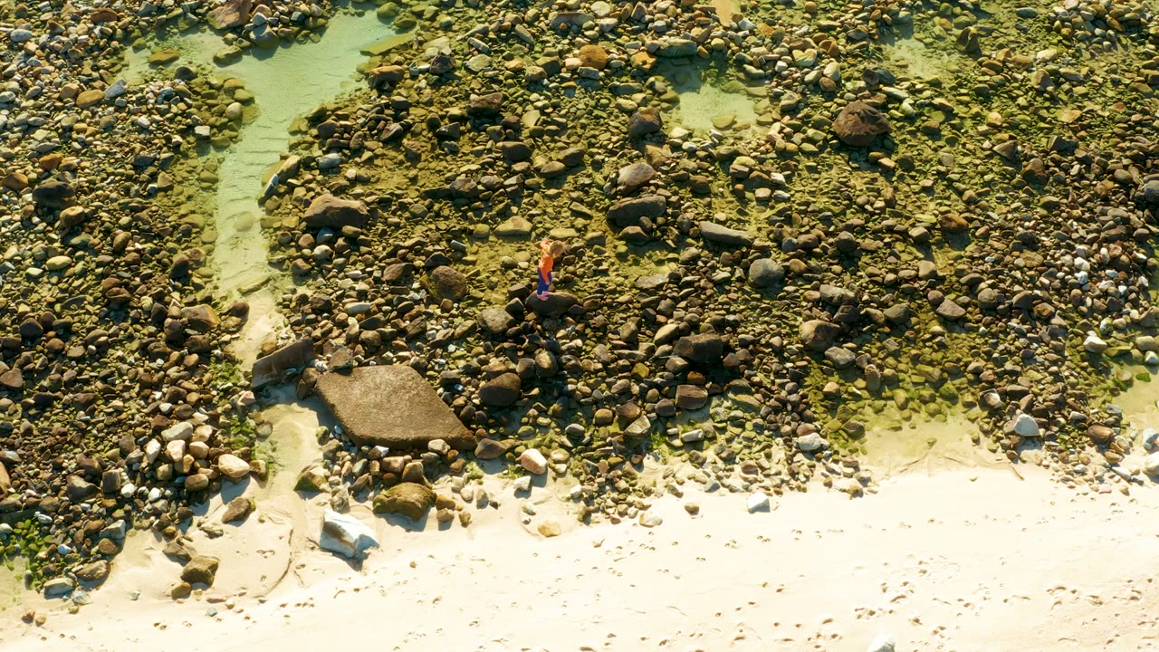 Overhead frame from El Tule Los Cabos with the person in red set against dense rocks, a pale water channel at left, and sunlit sand at the lower edge.