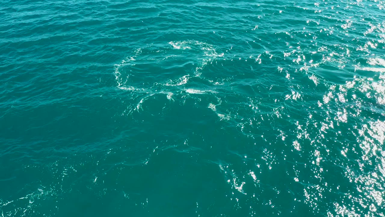 Turquoise ocean surface at El Tule Los Cabos with small ripples, scattered white foam, bright sun glints to the right, and no visible people.
