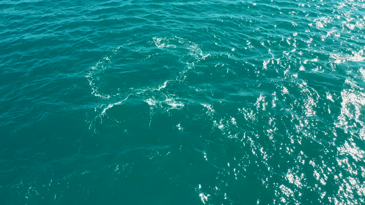Turquoise water at El Tule Los Cabos shows scattered white foam, fine ripples, a dense patch of sun sparkle along the right side, and no visible people.