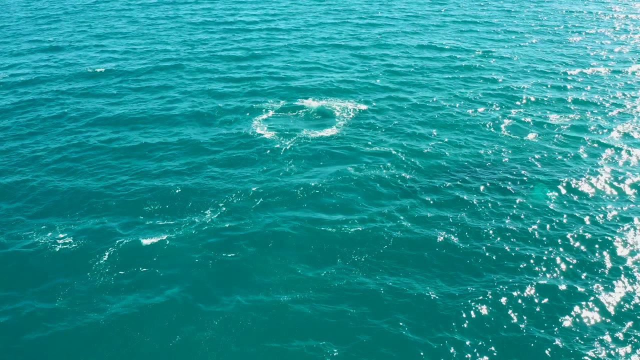 A compact white splash ring sits high in the frame over turquoise water at El Tule Los Cabos, with a strong vertical band of sun sparkle on the right and no visible people.