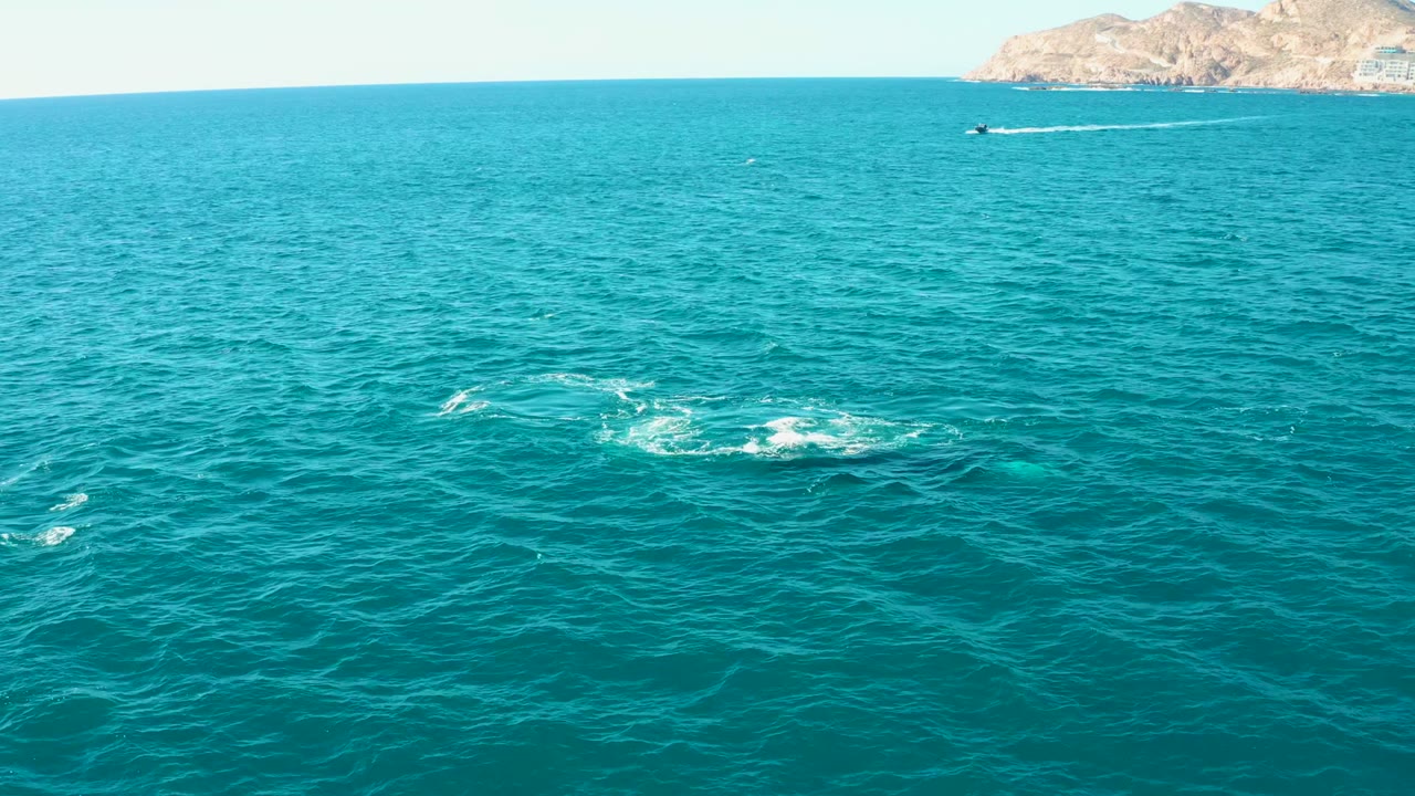Opening aerial view over turquoise ocean at El Tule Los Cabos, with several white splash rings on the water, small boats near the horizon, distant rocky land, and no visible people.