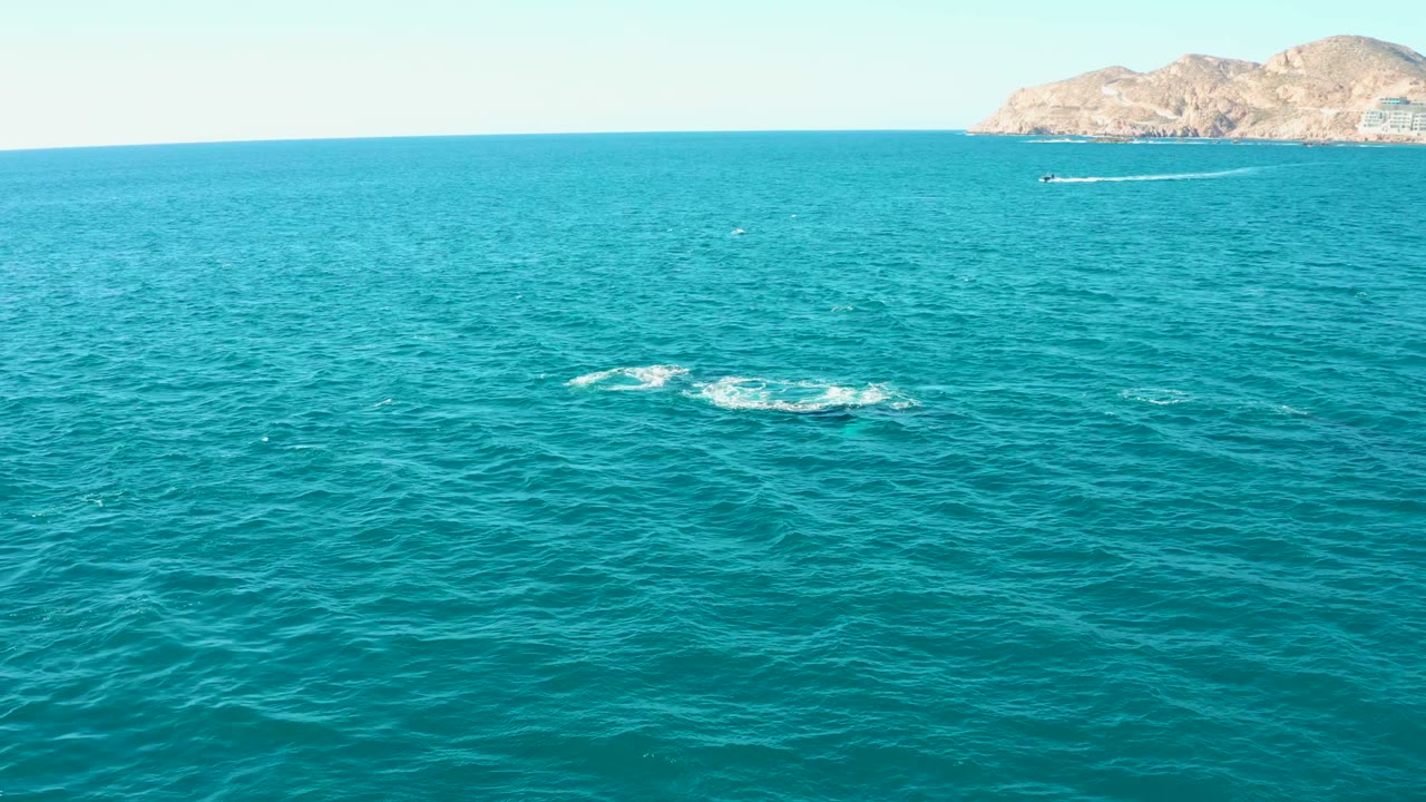 Aerial view over turquoise water at El Tule Los Cabos, with a small white splash in the midground, distant rocky land near the horizon, a small boat, and no visible people.