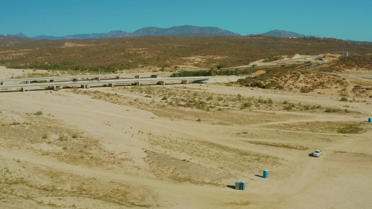 At 0:00 in El Tule, Los Cabos, a white car sits low in the sandy foreground while a highway bridge, scrub-covered hills, distant mountains, and clear sky fill the background, with no visible people.