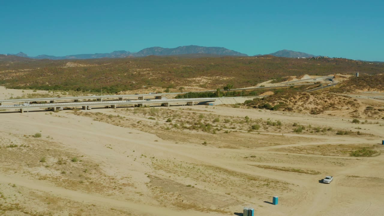 At 0:02 in El Tule, Los Cabos, the white car appears near the lower right edge of the sandy road area, backed by sparse scrub, blue markers, a highway bridge, hills, mountains, and no visible people.