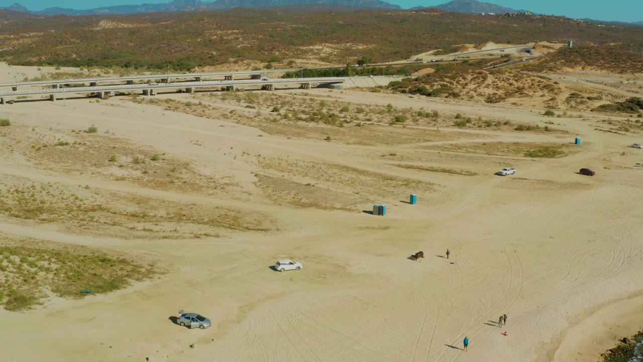 Aerial frame from El Tule, Los Cabos showing sandy off-road tracks, several cars, blue course markers, small visible people, a bridge, and dry hills beyond.