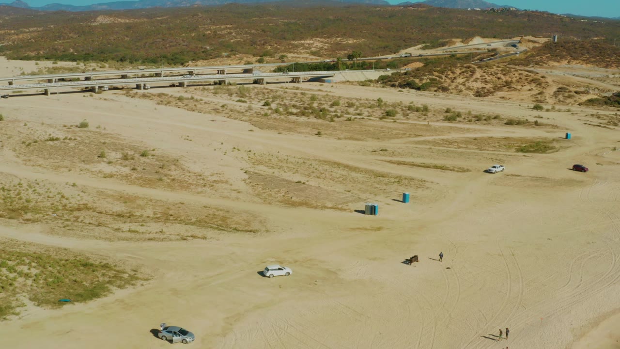 Wide aerial view of sandy course with cars, people, blue markers, bridge, and hills.
