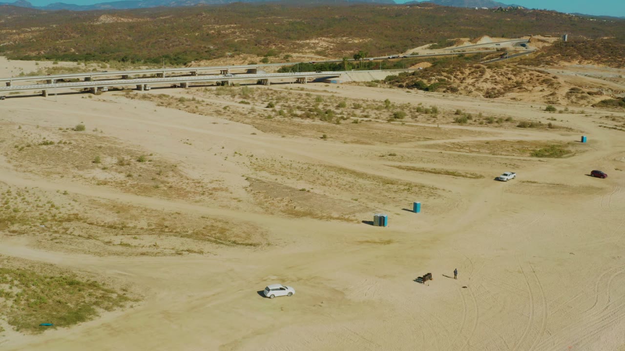 El Tule, Los Cabos aerial frame with a white car on sandy tracks, another dark vehicle nearby, small visible people near the course, blue markers, bridge structure, and distant mountains.
