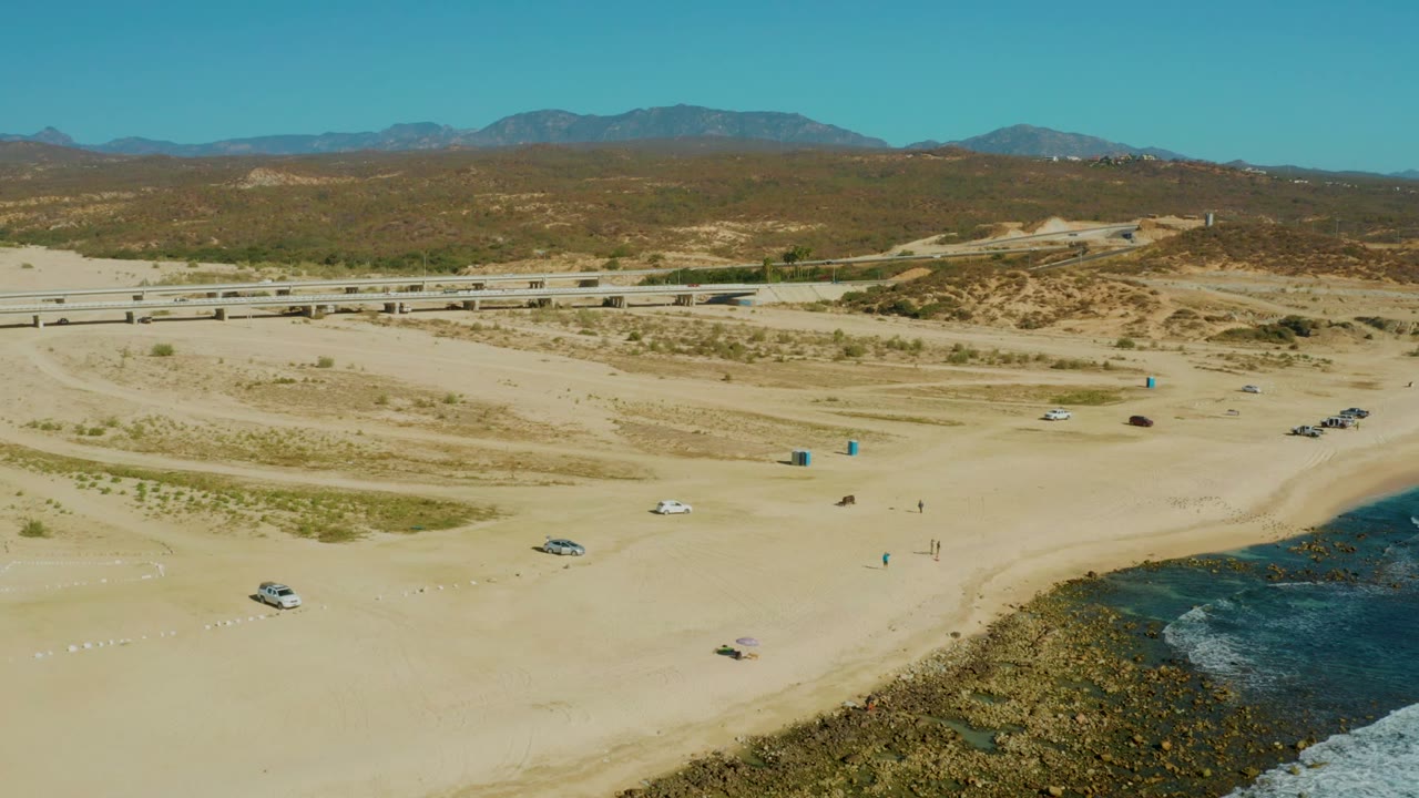 High aerial frame over El Tule Los Cabos showing sandy beach with scattered vehicles, small distant people, surf at the lower right, a road beyond the dunes, and mountains under clear sky.