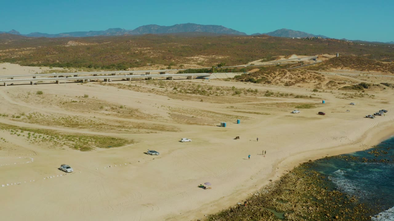 Wide drone view of beach activity with cars, people, road, hills, and shoreline.
