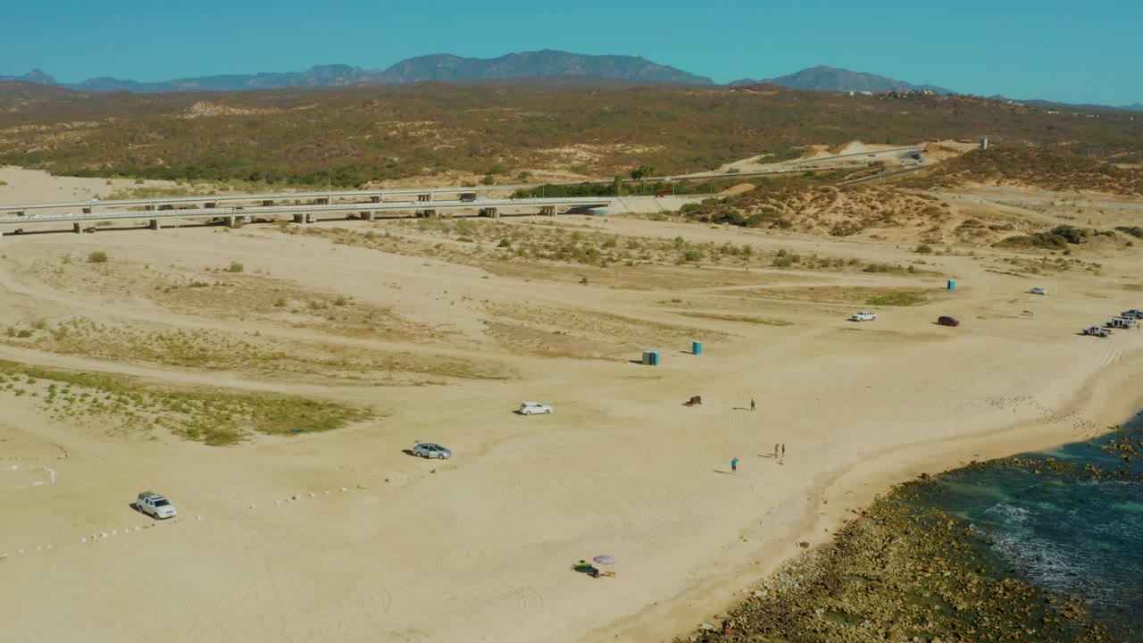 El Tule Los Cabos aerial frame with a white vehicle near the open sand, small people near the shoreline area, blue portable toilets near the center, waves on the right, and desert hills beyond the road.