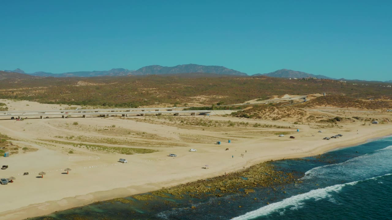 Aerial view over El Tule Los Cabos with sandy beach, rocky surf at the lower right, a coastal road across the middle distance, scattered vehicles, dunes, mountains, clear sky, and no visible people.
