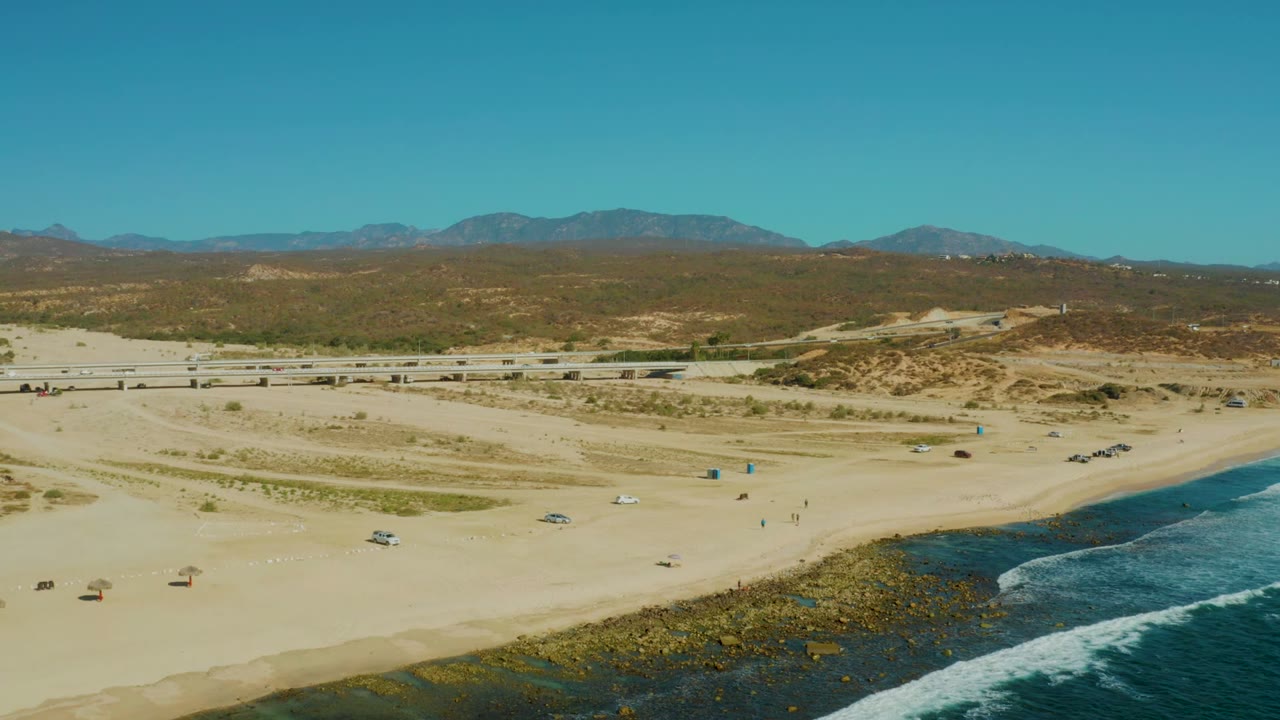 Wide coastal drone view with sand, road traffic, waves, dunes, and mountains.