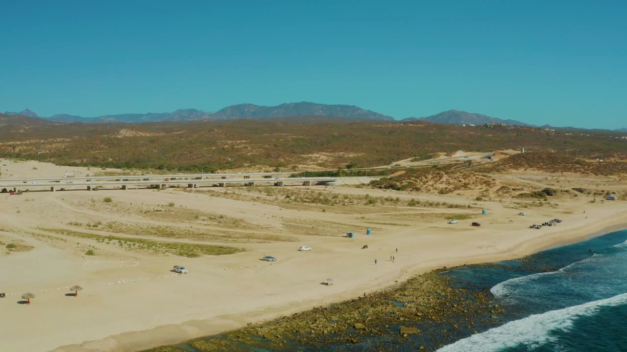 El Tule Los Cabos coastline from above with a broad sandy beach, cars spaced along the road and beach area, white surf crossing the rocky edge, desert dunes behind the road, mountains on the horizon, and no visible people.