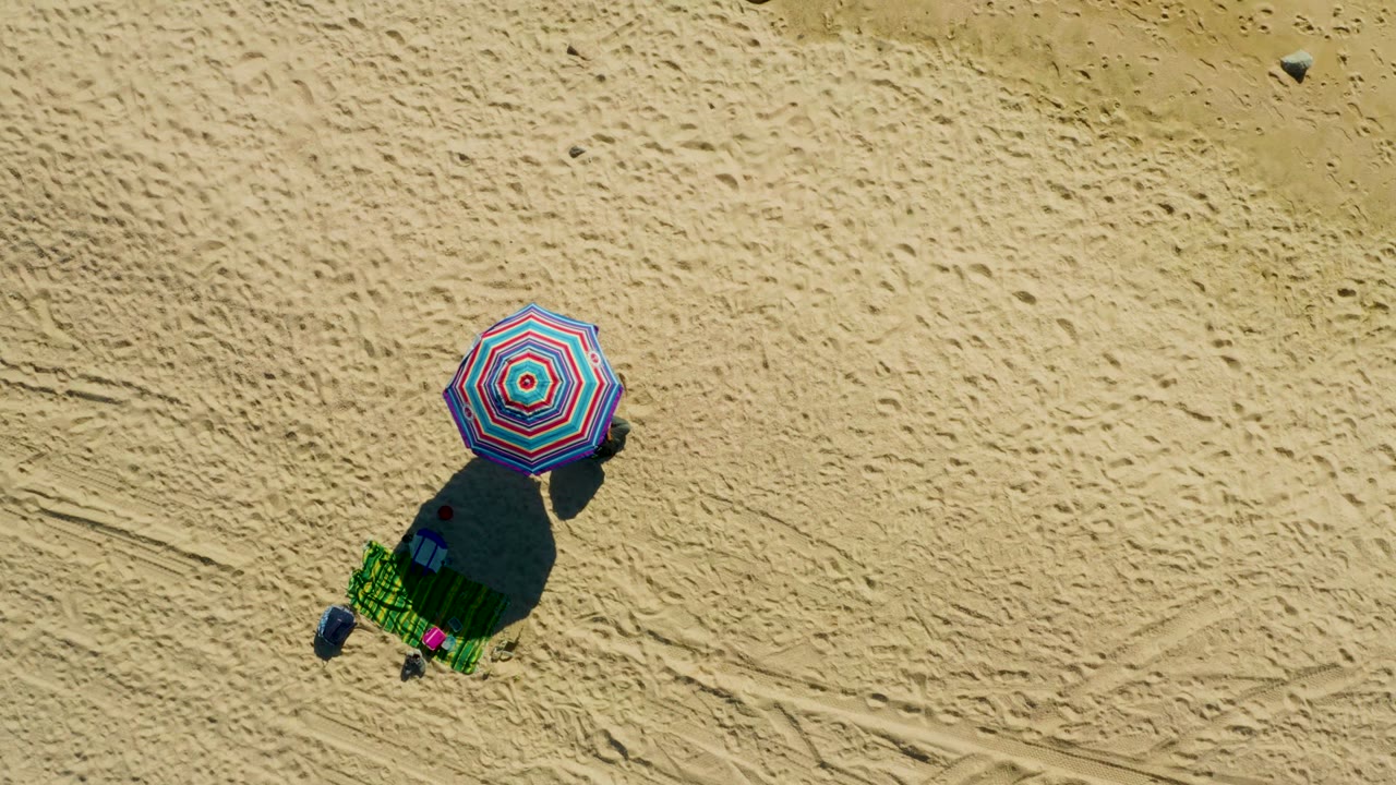 At El Tule Los Cabos, an overhead view shows a striped beach umbrella, green lounge chair, small items, and a tiny visible person on marked sand at the start of the clip.