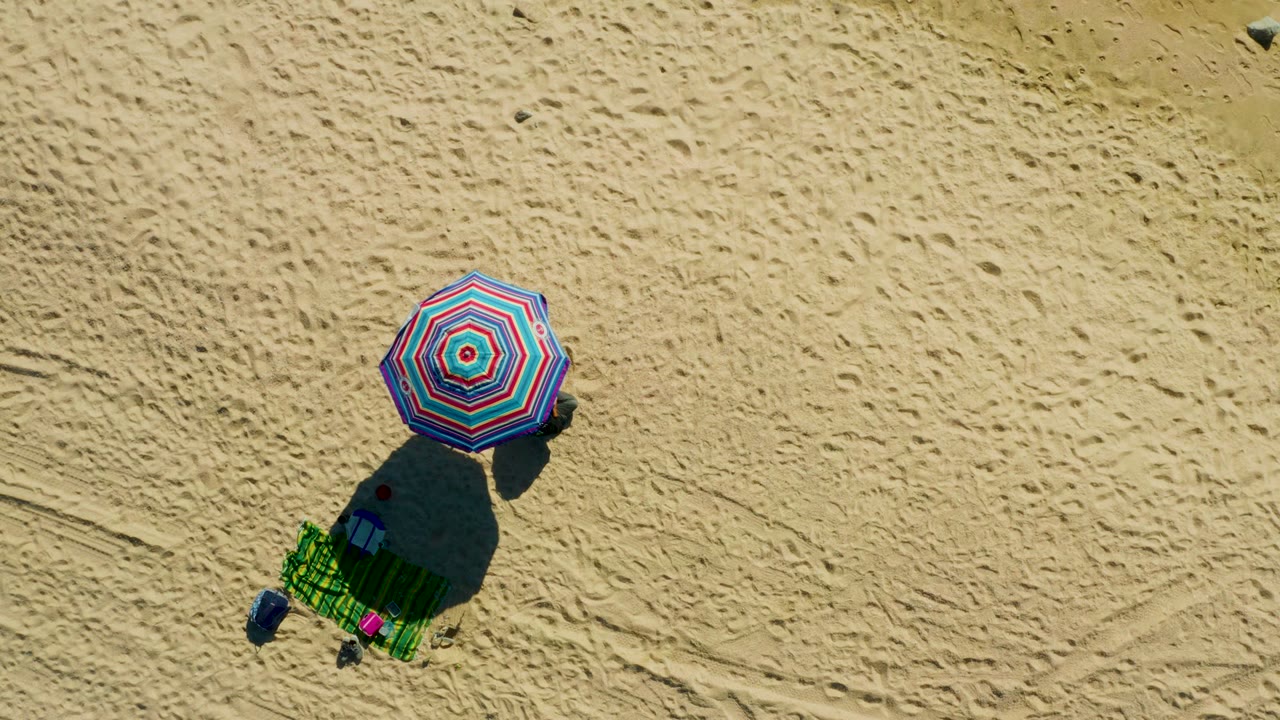 At El Tule Los Cabos, the top-down frame shows the circular umbrella pattern, green chair, dark cast shadow, scattered small items, and textured sand around the beach setup.