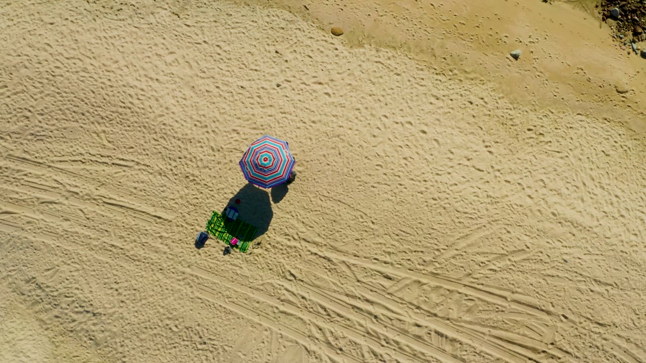 Overhead view at El Tule Los Cabos shows a striped umbrella, green chair, beach items, long shadow, tire tracks, and no visible people.