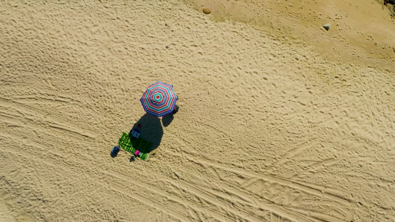 At 0:02, the overhead El Tule Los Cabos view shows the umbrella and chair over pale sand, with visible track patterns and no visible people.