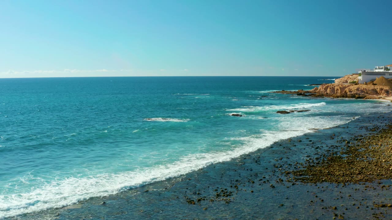 Wide elevated view of El Tule Los Cabos turquoise ocean with white surf crossing a dark rocky shore, tan headland at the right edge, and no visible people.