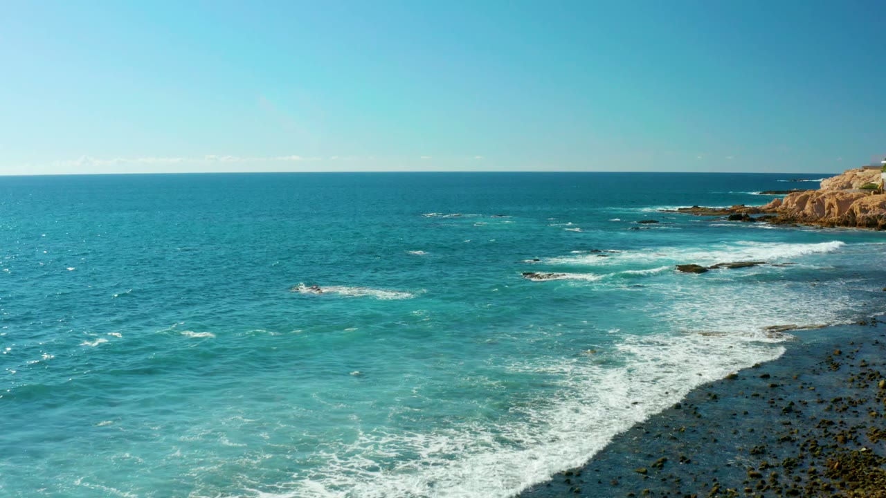 Wide El Tule Los Cabos ocean frame with deep blue water offshore, turquoise water near the rocks, white foam curling along the shore, and no visible people.
