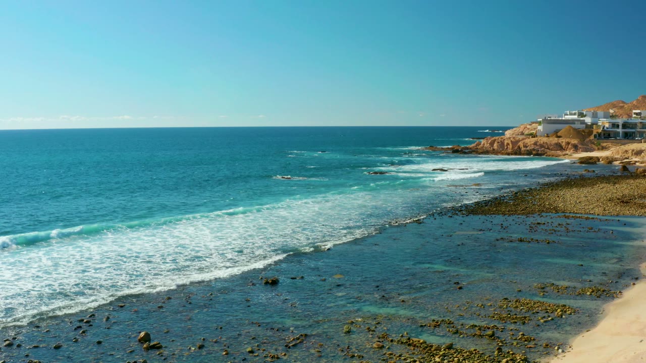 At 0:00 in El Tule Los Cabos, blue water and white surf spread across a rocky shoreline below white cliffside buildings, with no visible people.